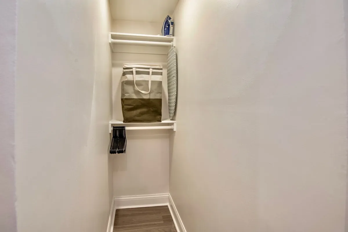 A practical bedroom corner with an ironing board, iron, hangers, and a tote bag. The organized setup emphasizes the room's ample storage options and the ability to keep clothing tidy and ready, underscoring the space's suitability for longer stays or life transitions.