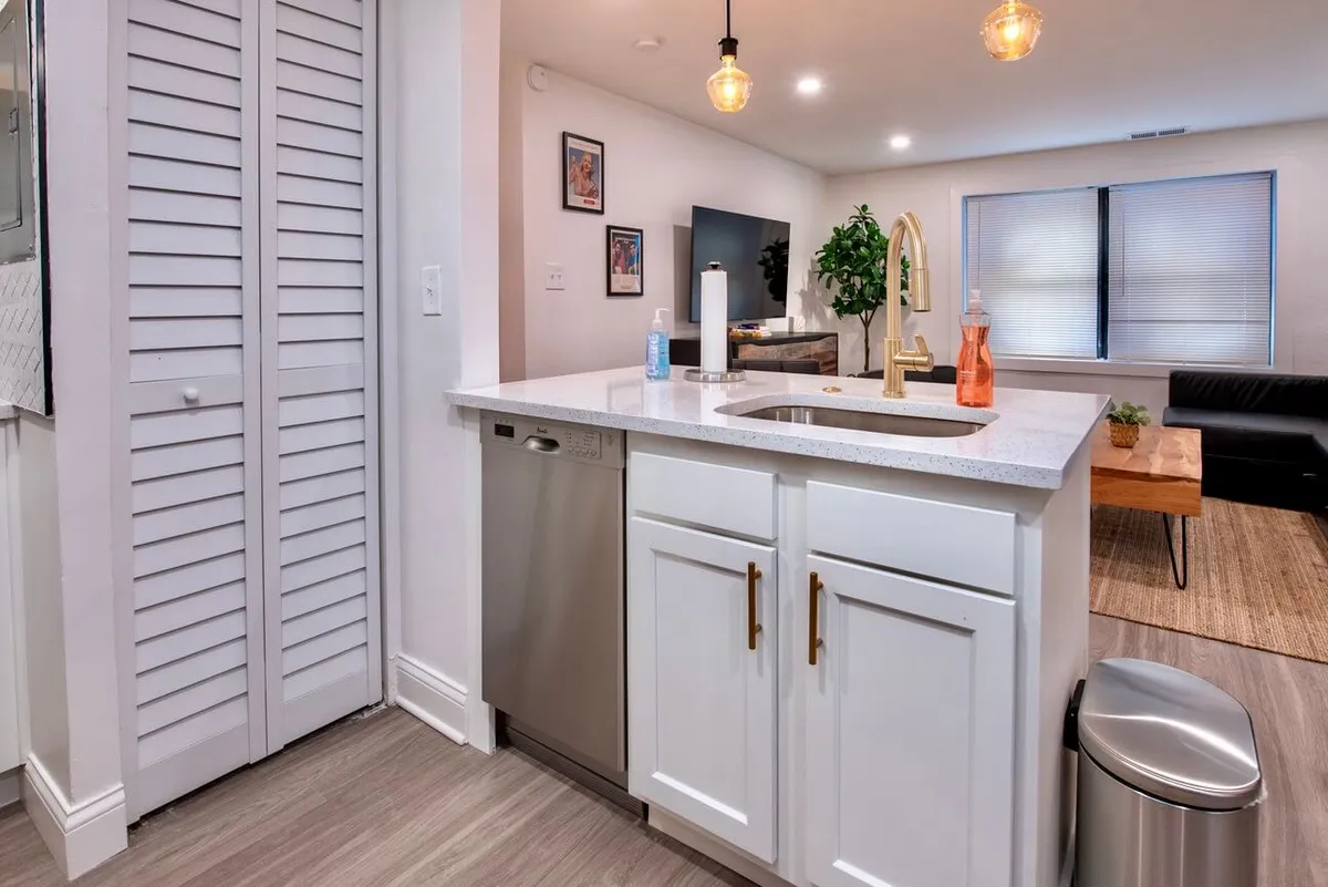 Showing another angle of the kitchen, this photo highlights a kitchen sink, integrated dishwasher, golden pendant lights, a touch of greenery with a houseplant, and framed artwork. The cheerful and contemporary mood reinforces the kitchen’s inviting atmosphere, while also illustrating functional features.