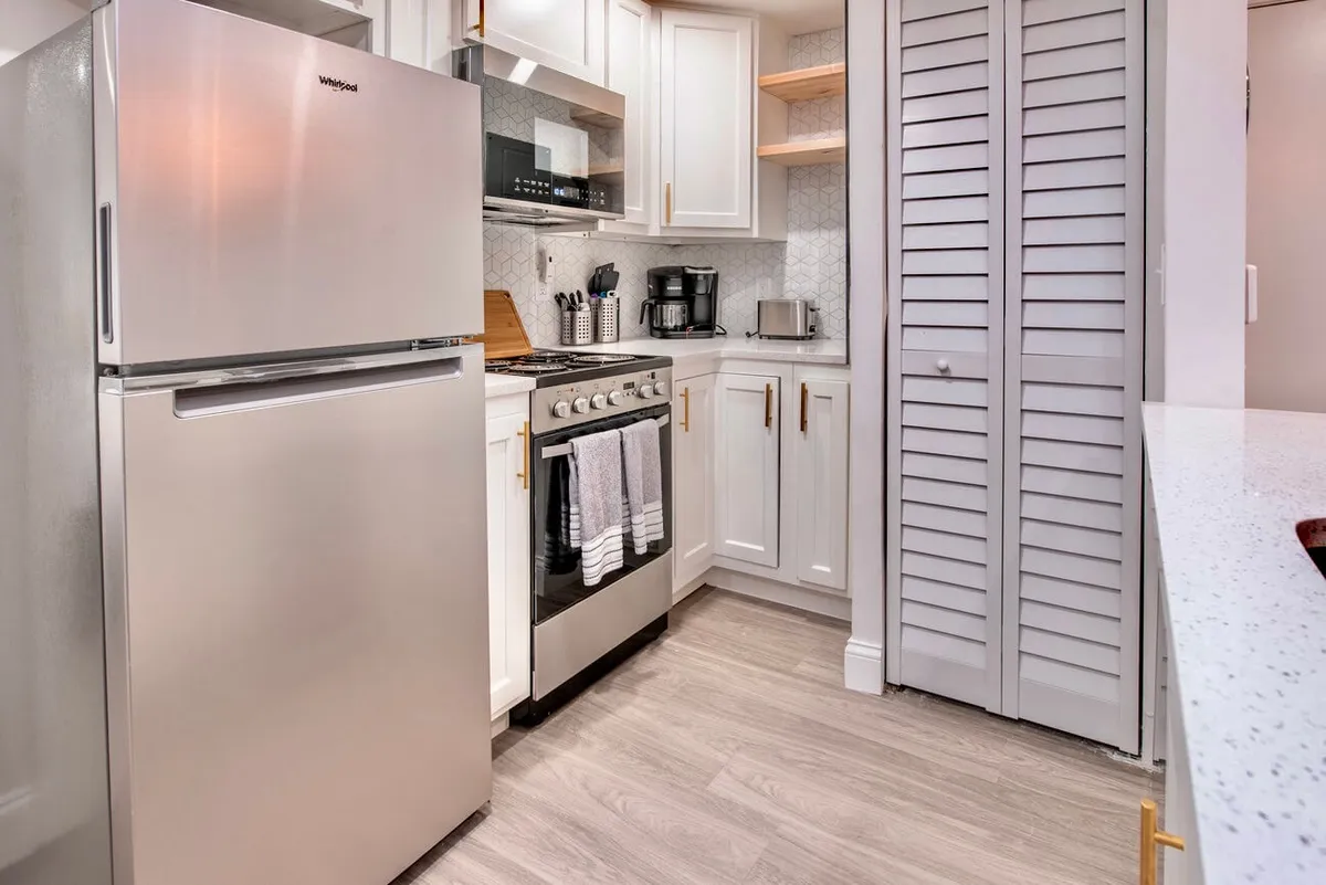 This image captures the heart of the kitchen, featuring a stainless steel refrigerator, a modern stove, crisp white cabinetry, and stylish open shelving. The scene suggests both functionality and a welcoming space for meal preparation or casual gatherings, with ample counter space visible for everyday cooking tasks.