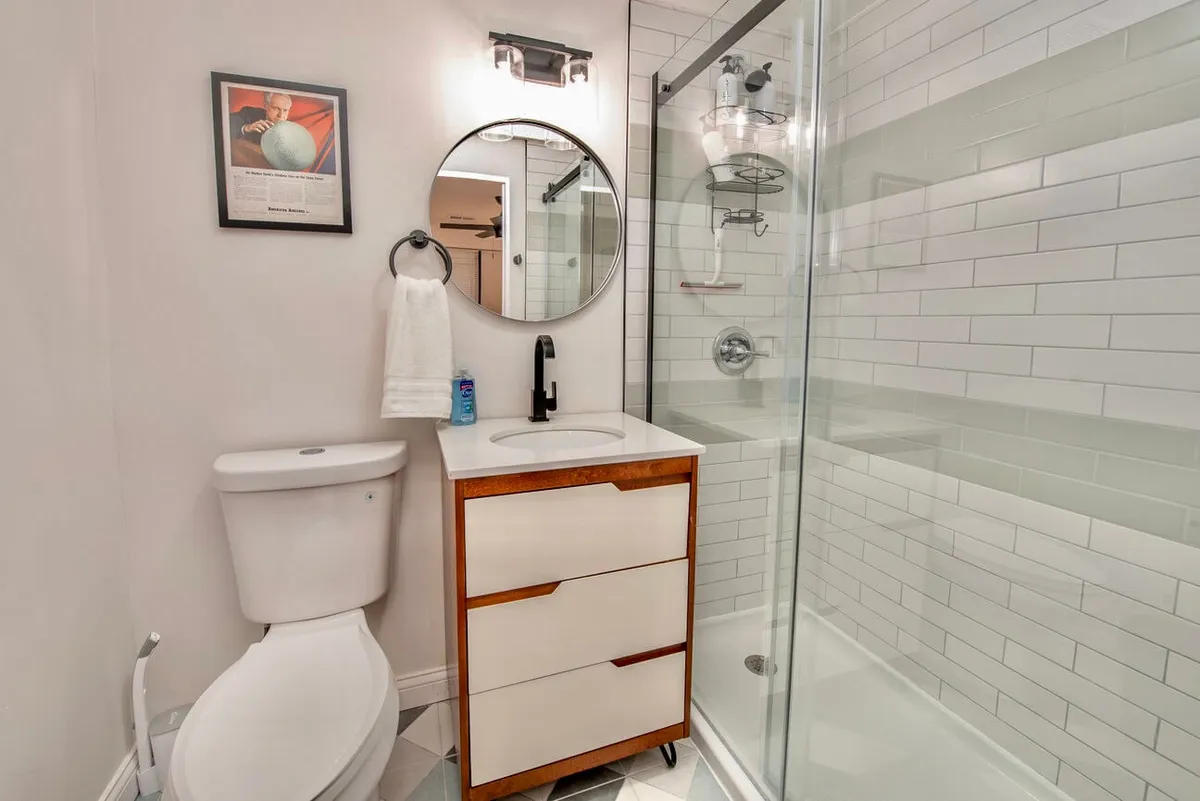 This image showcases a modern bathroom featuring a sleek wooden vanity, a round mirror, and a glass-enclosed shower. The finishes are clean and contemporary, with bright lighting that highlights the fresh and inviting atmosphere. Subtle details like organized toiletries and fresh towels suggest both comfort and functionality.