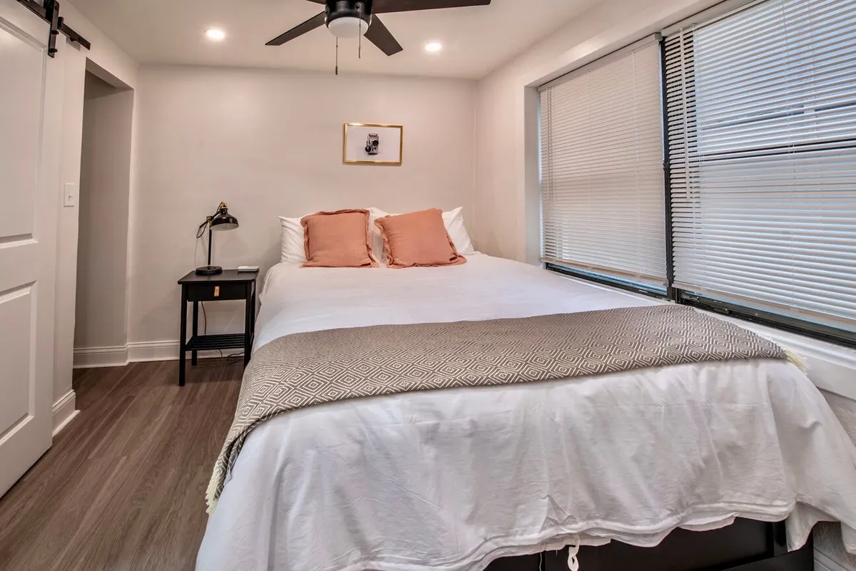 A cozy bedroom featuring a neatly made bed adorned with plush decorative pillows. Soft natural light filters in through window blinds, while a desk lamp and framed artwork contribute to the tranquil atmosphere. Every element, from the bedding to the decor, reinforces a sense of comfort and relaxation.